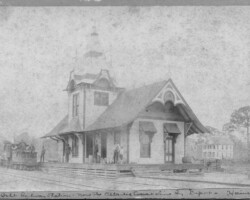 The Orange Belt Railway depot, which once stood at the corner of 1st Ave S and 2rd St. A narrow gauge engine is to the left of the platform, and the end of a boxcar is visible to the right. Four men and a boy stand on the platform. Orange Belt Investment Company office is in the background. c. 1889